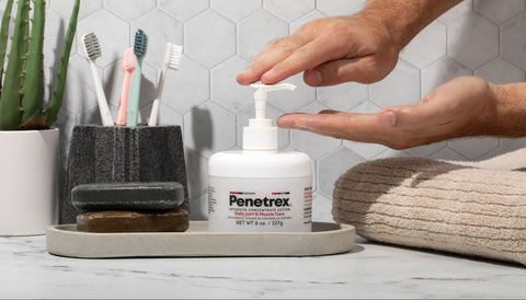 Person applying Penetrex cream from a pump bottle on a bathroom counter with toothbrushes and a plant in the background.